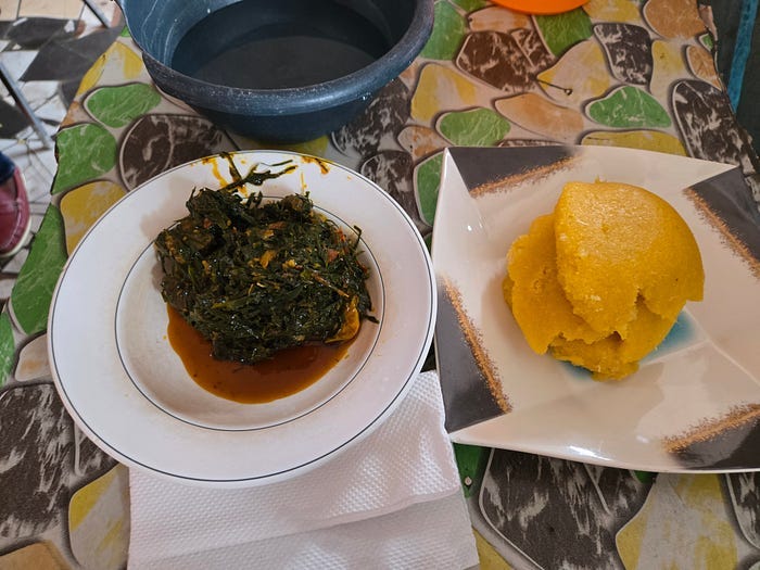 A green ball of leaves and meat on a plate, next to a pancake-like patty of fufu. A green ball of leaves and meat on a plate, next to a pancake-like patty of fufu.