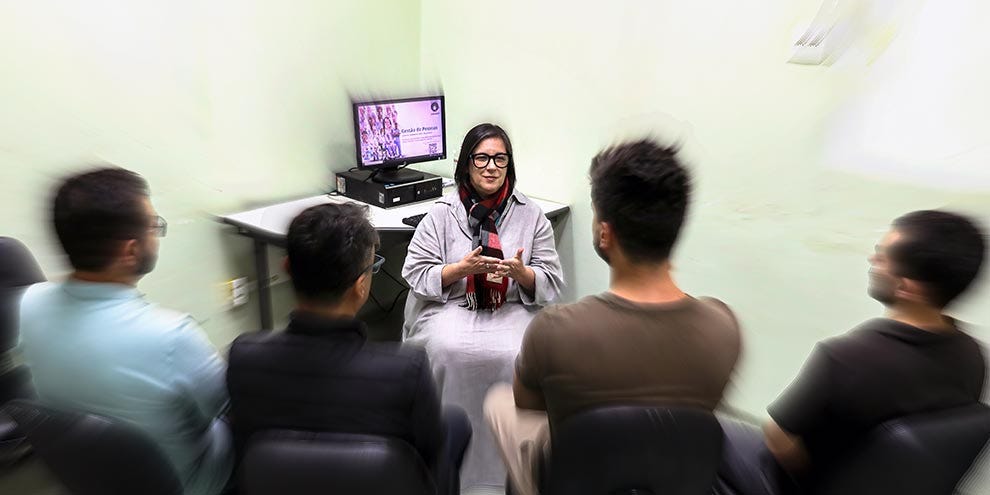 Pessoa ao centro usando óculos, camisa clara e colar vermelho, circundada por quatro pessoas de costas vestindo camisetas em tons de azul, preto e marrom, em sala com parede clara, mesa com monitor de televisão ao fundo exibindo conteúdo em roxo e equipamento eletrônico preto. Pessoa ao centro usando óculos, camisa clara e colar vermelho, circundada por quatro pessoas de costas vestindo camisetas em tons de azul, preto e marrom, em sala com parede clara, mesa com monitor de televisão ao fundo exibindo conteúdo em roxo e equipamento eletrônico preto.