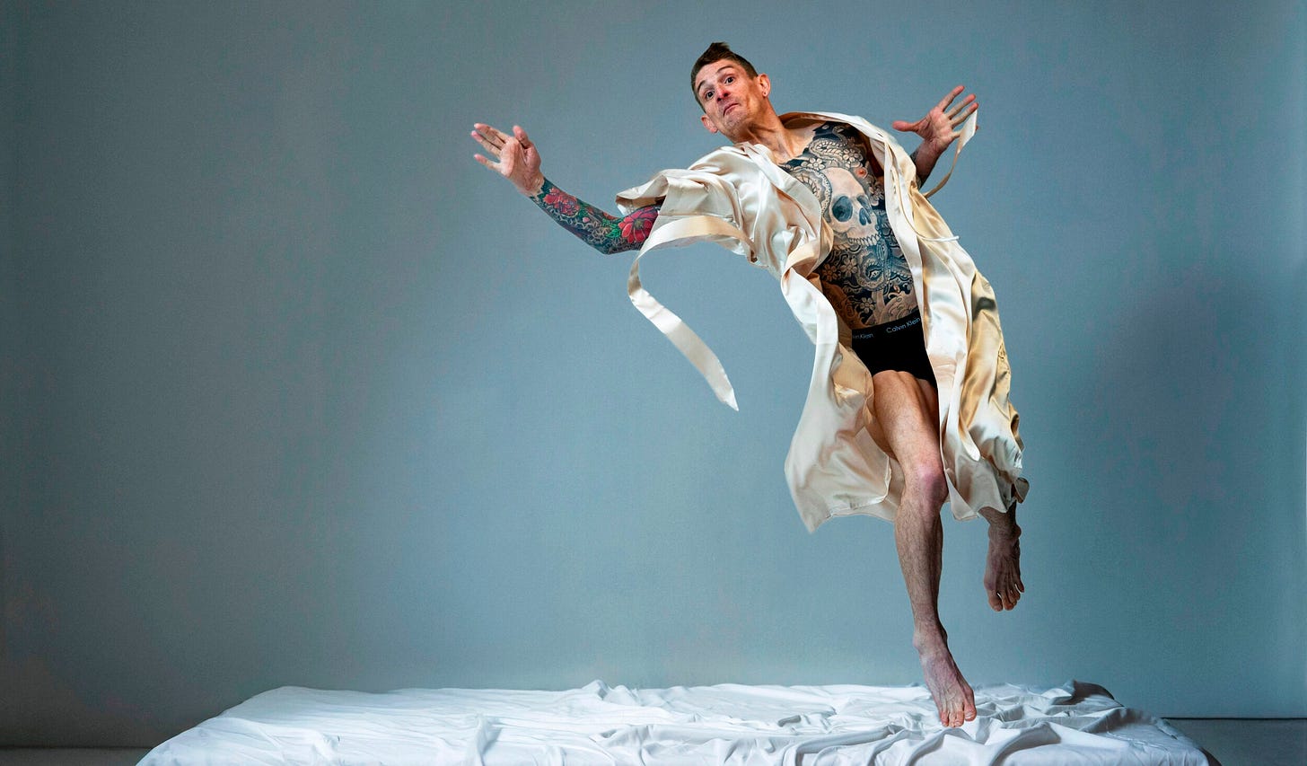 Dan is a tattooed man who is airborne and reaching for something. He is wearing black briefs and a cream silk dressing gown set against a grey background.
