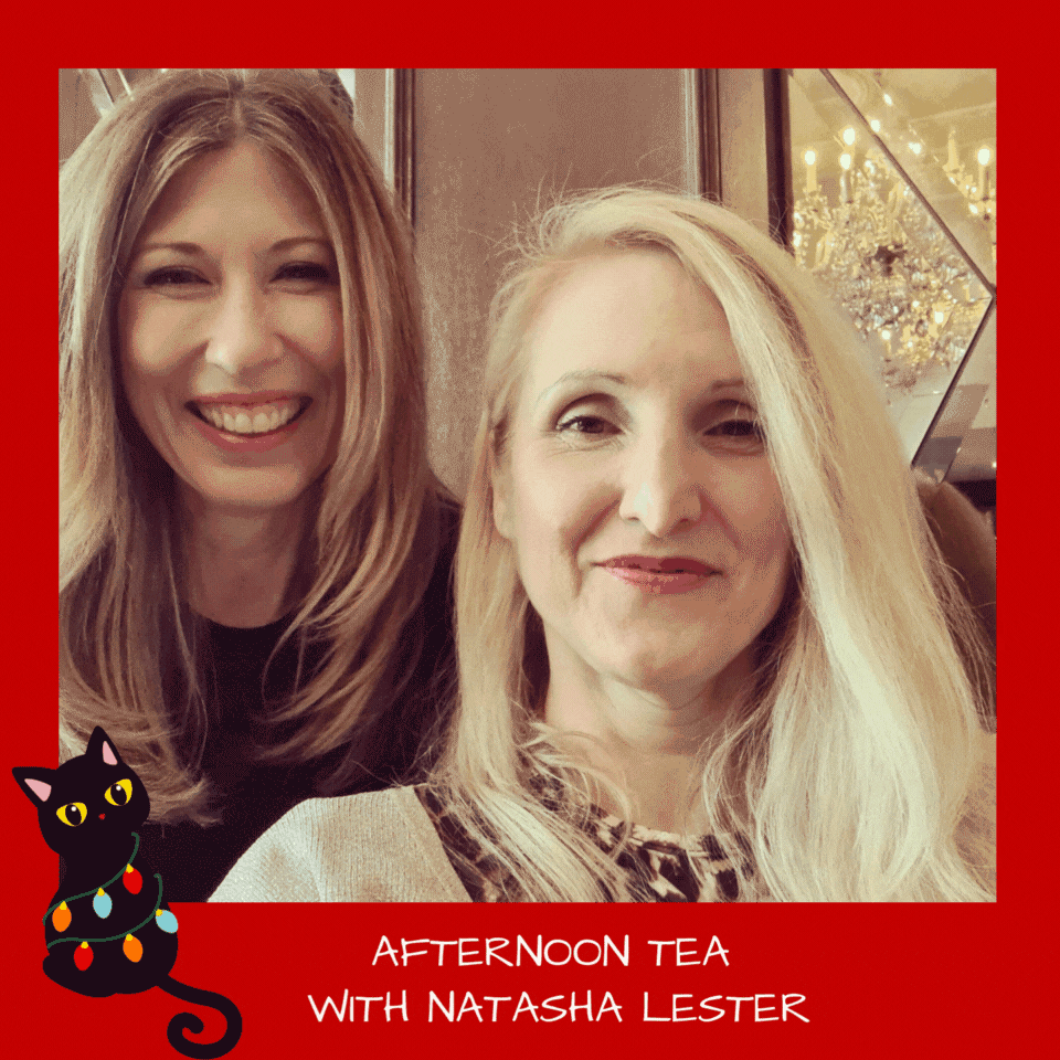 A close up selfie of two women having an excellent time together. The woman on the left with dark blonde hair and a big smile is Natasha Lester. On the right with blonde hair and a big smile is Belinda Alexandra. The image is surrounded by a bright Red border and has a black cat cartoon wearing coloured lights.