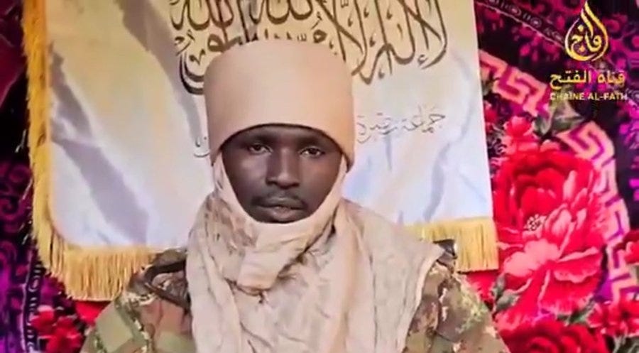 A man with dark skin wearing a light beige turban and wrapped headscarf stands in front of a large green banner with white Arabic script and a logo resembling a sword or emblem. He is dressed in camouflage military attire over his shoulders and a light scarf around his neck. The background includes decorative pink and purple fabric elements.