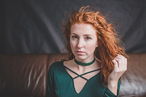 Woman, model, red wavy hair, freckles, green top and choker, pensive expression, sits on leather sofa