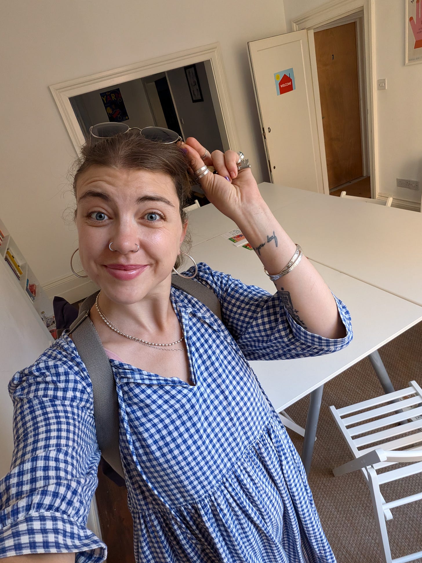 Grace in her Writing Workshop space, wearing a blue and white gingham dress, sunnies on her head, posing with the room and smiling inoffensively at the camera.
