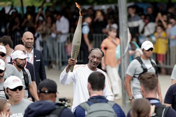 Snoop Dogg, in a white track suit, carrying the Olympic torch through a crowd of onlookers in Paris.