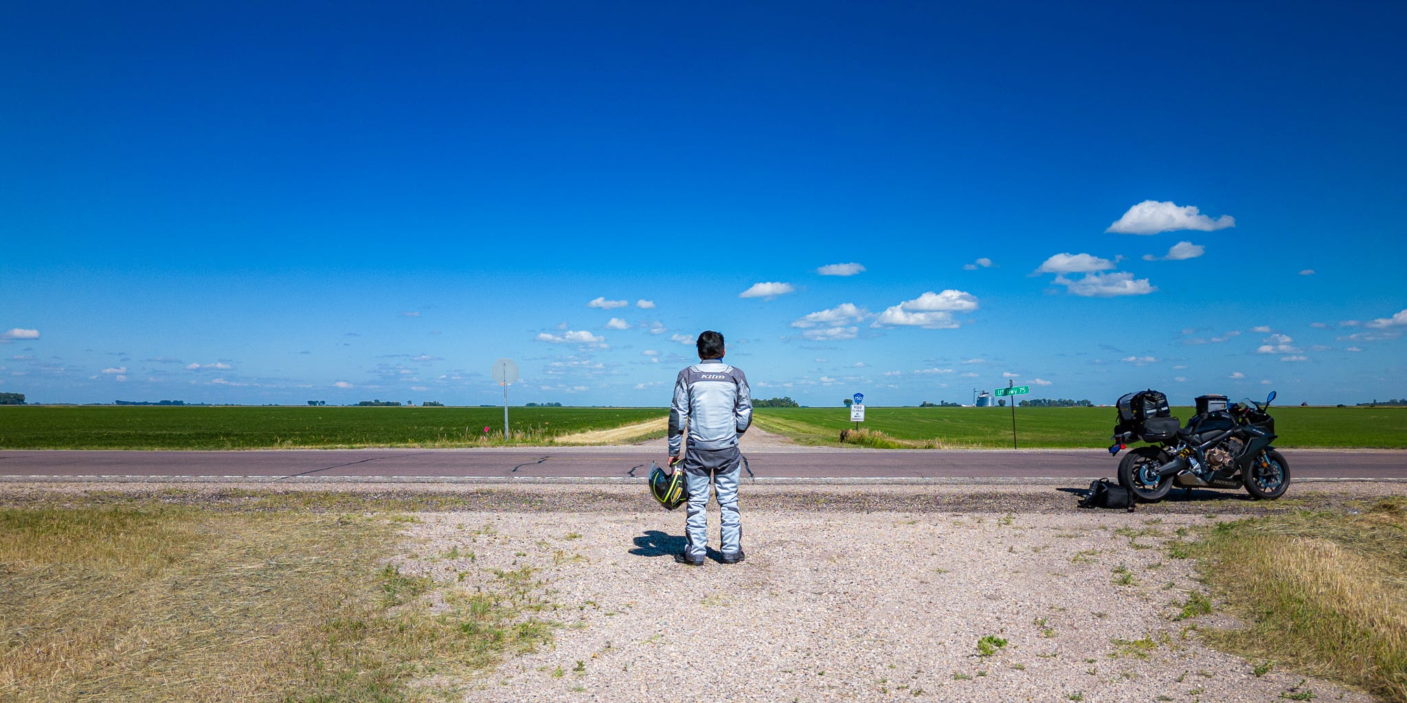 photo of a man in motorcycle riding gear on the side of a empty farm road. His motorcycle is parked on the right along the side of the road. The man is looking across the road, towards an empty gravel farmroad pointing towards the horizon. On either side of the road are green fields. The sky is blue and dotted with clouds