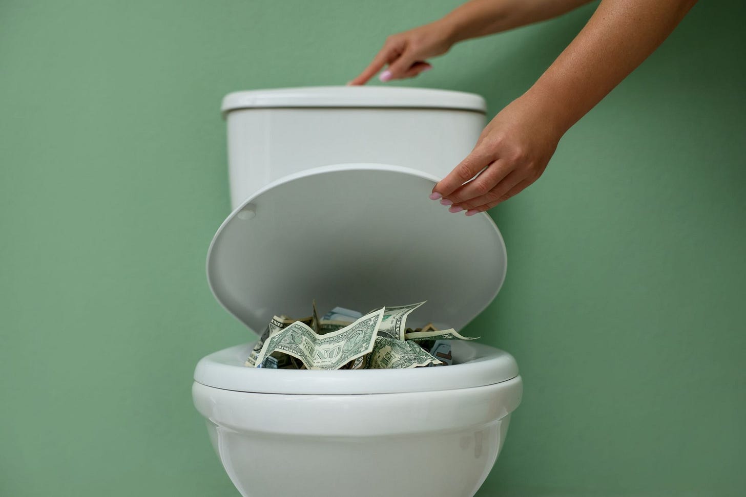 White porcelain toilet full of bank notes against a green wall. The person closing the toilet seat lid is about to press the flush on top of the back of the toilet with their finger, and flush all the money away.