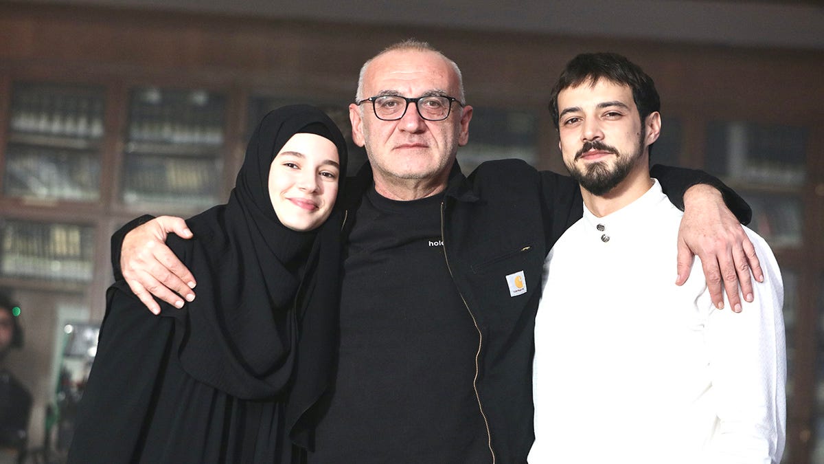 Üç kişi, bir kapalı mekanda samimi bir şekilde poz veriyor. Soldaki kişi siyah çarşaf giyerken, sağdaki kişi beyaz bir kıyafet tercih etmiş. Arka planda kitaplarla dolu raflar bulunuyor.