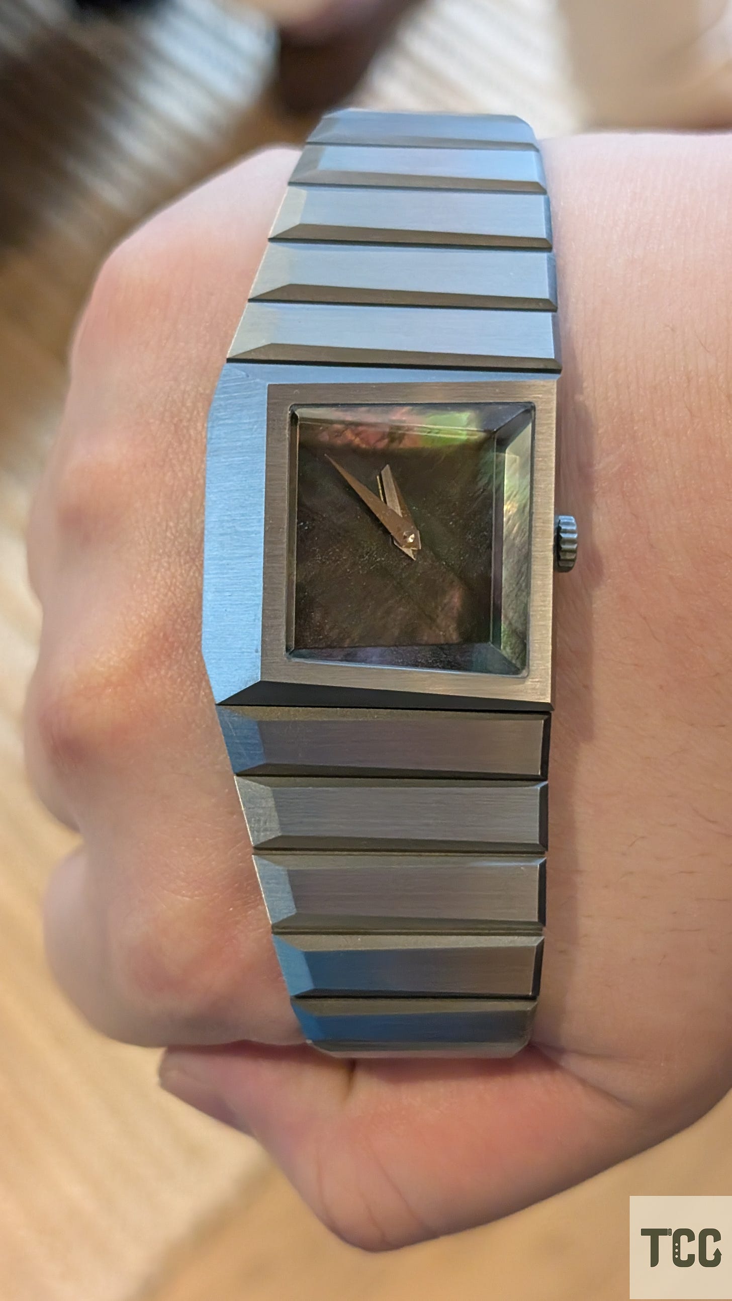 A close-up wrist shot of a futuristic, brutalist-style watch with a brushed metal bracelet and a square case. The dial appears to be black mother-of-pearl, reflecting iridescent greens and purples. No markers or brand name are visible.