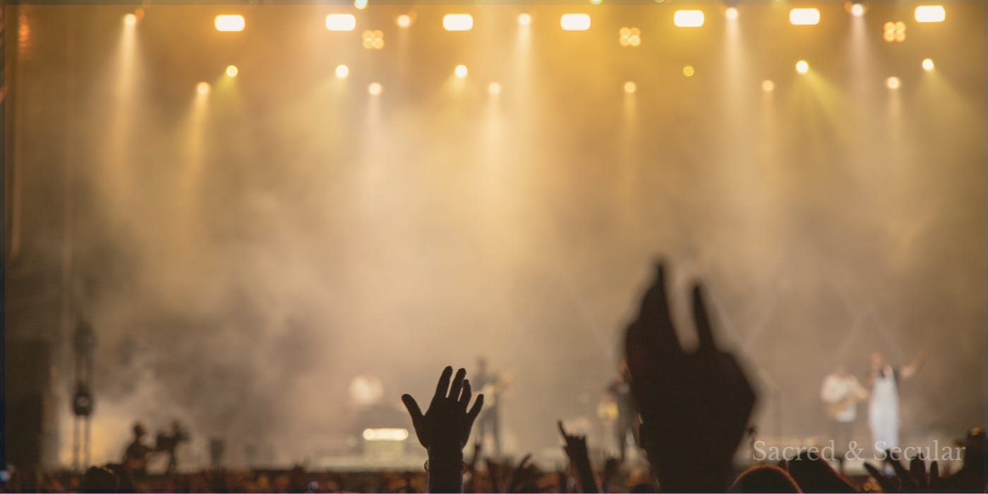 A concert stage illuminated by warm golden spotlights shines through haze as silhouetted audience members raise their hands in the foreground, capturing the atmosphere of a live music performance. A concert stage illuminated by warm golden spotlights shines through haze as silhouetted audience members raise their hands in the foreground, capturing the atmosphere of a live music performance.