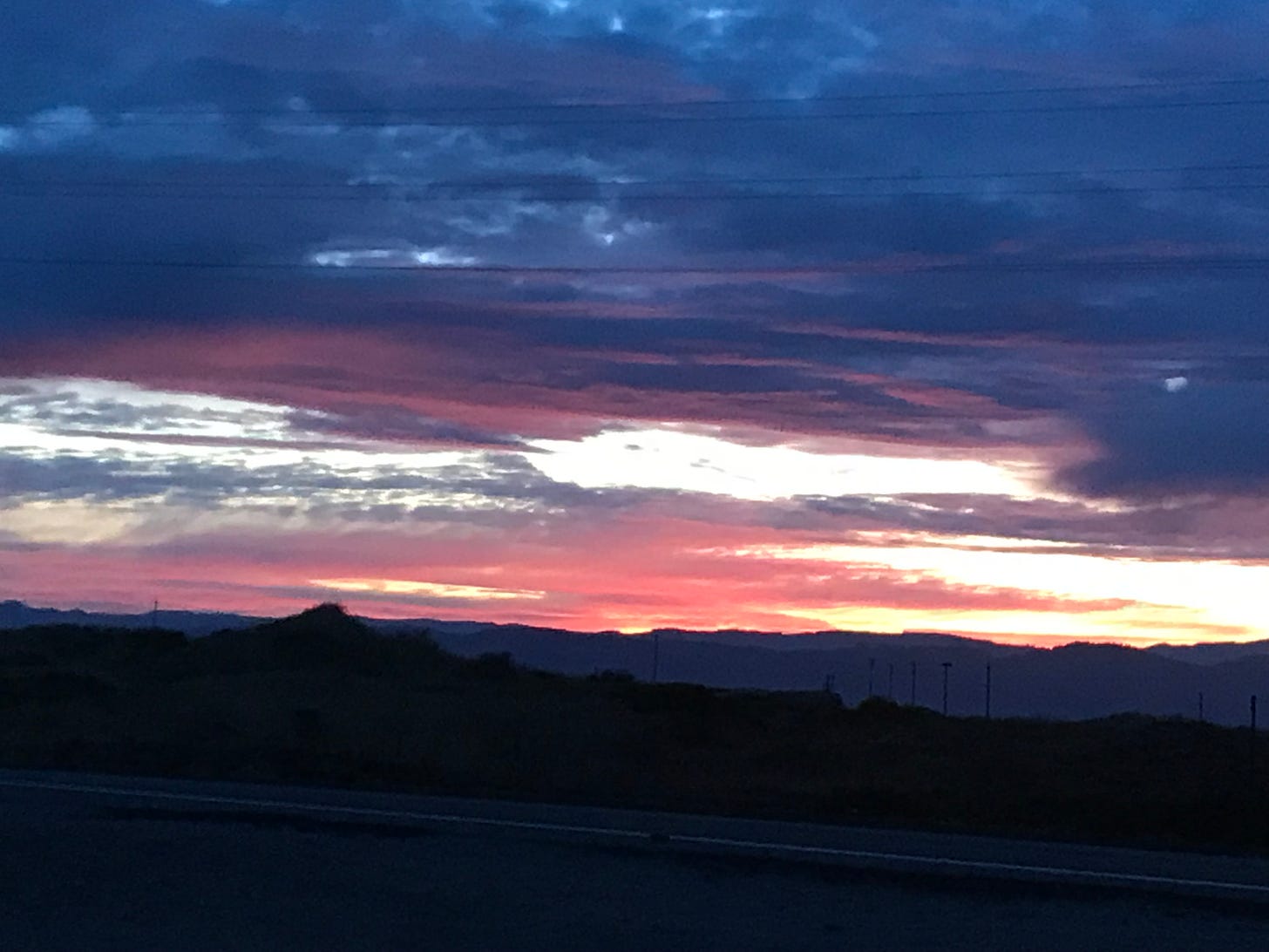 A dark blue cloudy sky over streaks of pink and peach and golden sunrise over navy sand dunes