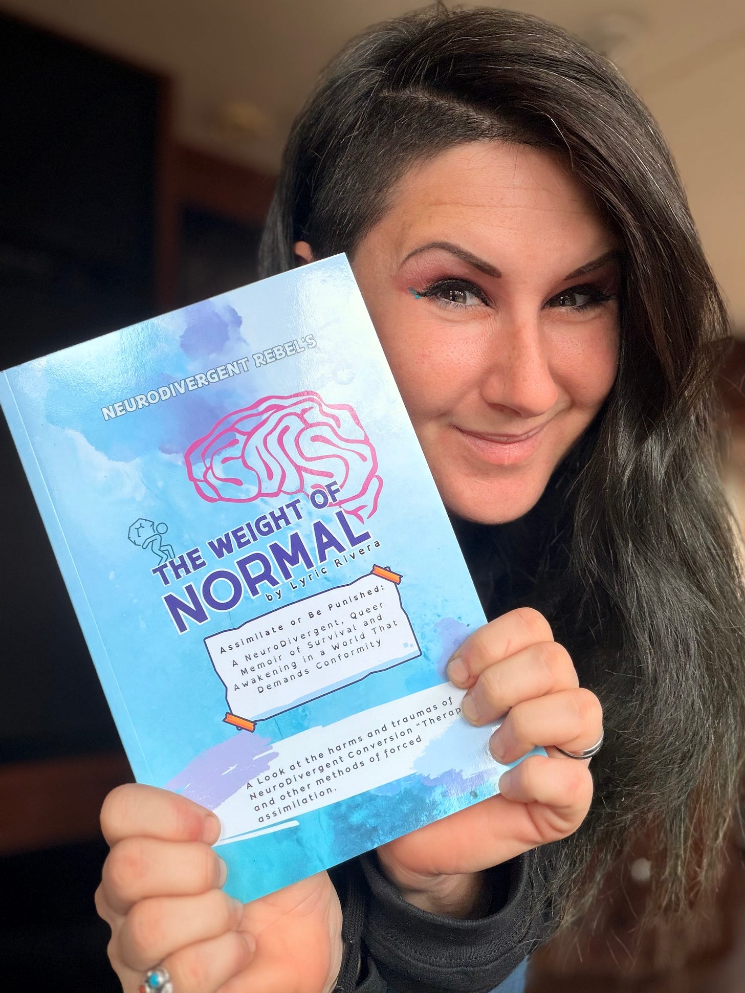 A happy Lyric, showing off their latest book, ‘Neurodivergent Rebel’s: The Weight of Normal’ - a teal book with a pink brain on the cover.