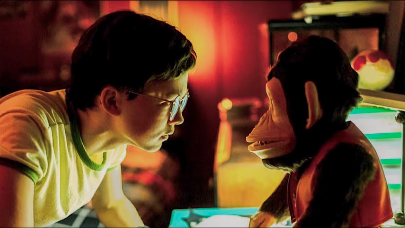 A young boy in glasses leans in, face-to-face with a grinning monkey toy, faces lit from underneath and backlighting.
