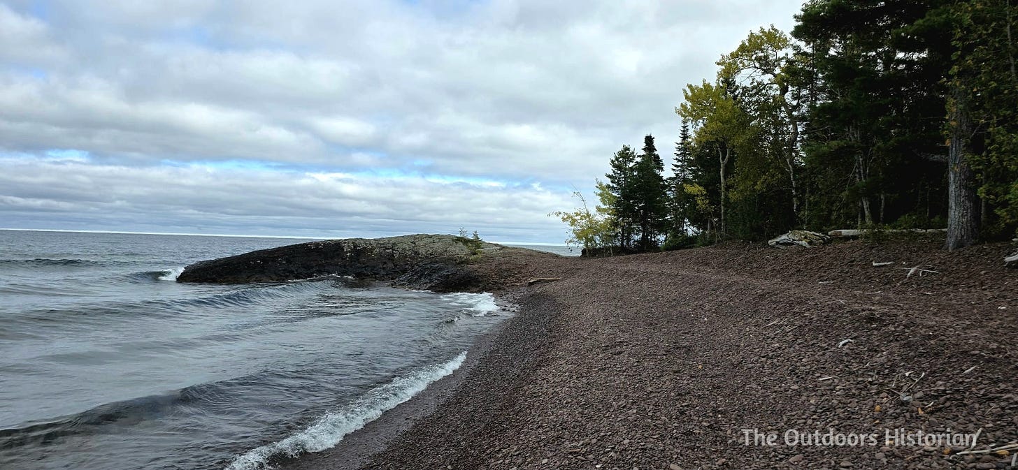 A rocky beach made up of mainly small cobble leading to a small basalt point. There a small waves on the lake and many fluffy clouds in the sky with the occasional sliver of blue.