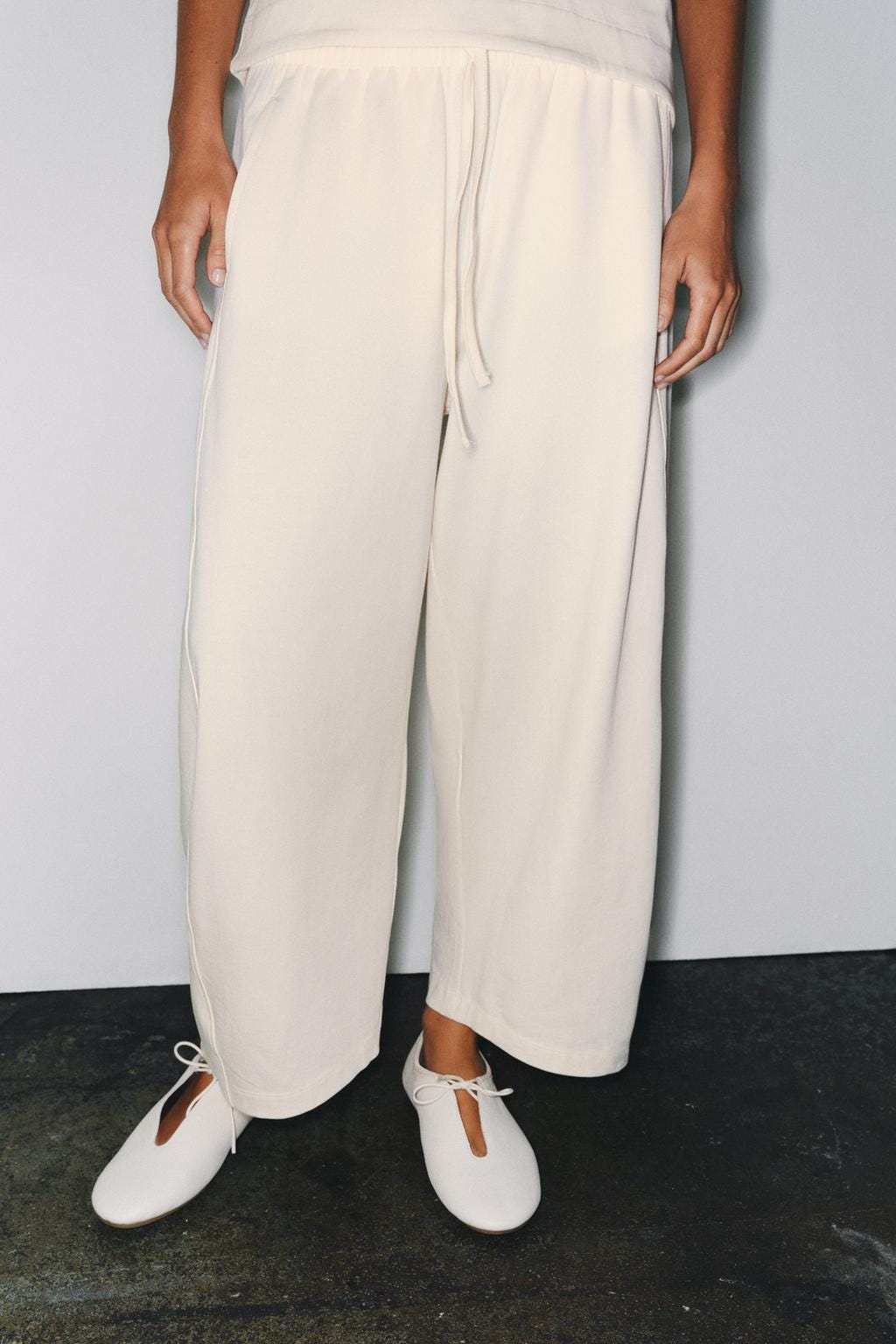 Mid-shot of ecru wide-leg trousers with an elasticated waist and a front tie. White flat shoes with lace detailing.