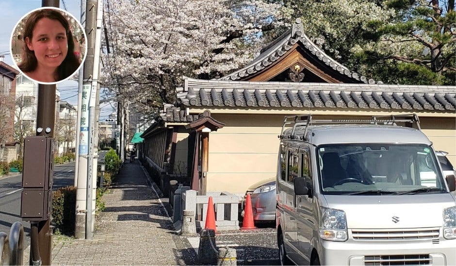 A quiet street in Japan with a narrow sidewalk running alongside a traditional temple wall topped with ornate roof tiles. Cherry blossom trees in full bloom rise above the wall, scattering petals onto the path. A silver Suzuki van is parked on the street beside orange traffic cones, and power lines run overhead. In the upper left corner, a circular inset shows a smiling woman with brown hair.