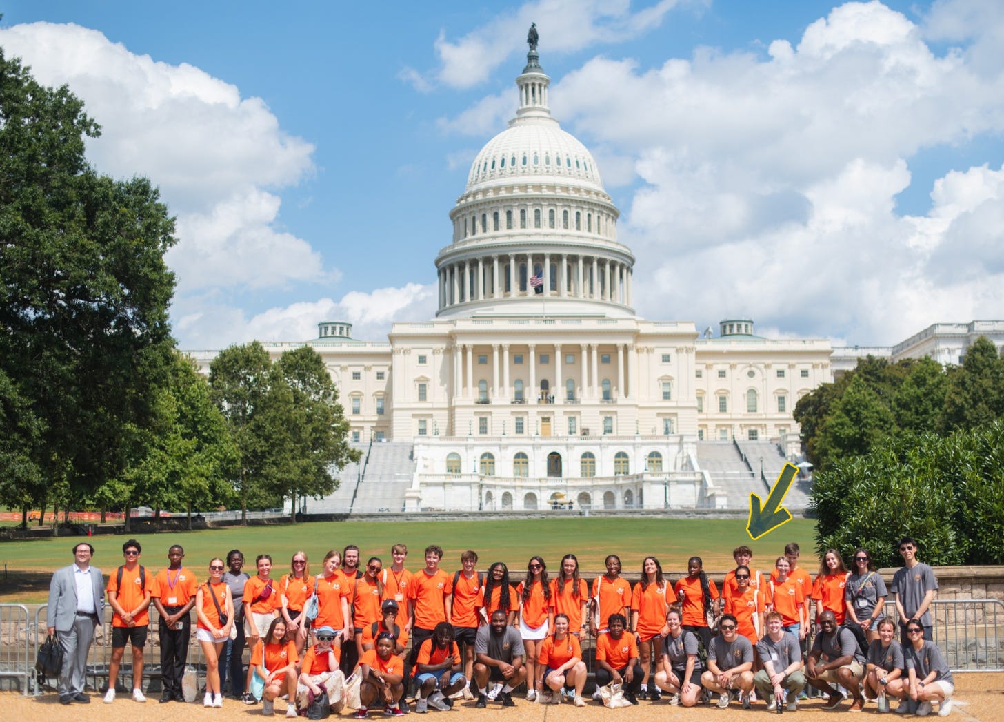 A large group of young people poses in front of the U.S. Capitol building on a sunny day. Most of the group is wearing bright orange T-shirts, while a few are in gray. William, near the right side of the group, is marked with a yellow arrow.