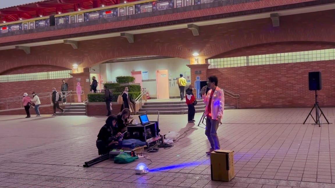 A man dressed in a white blazer wrapped in Christmas lights sings karaoke in the square outside Taipei's Tamsui MRT station
