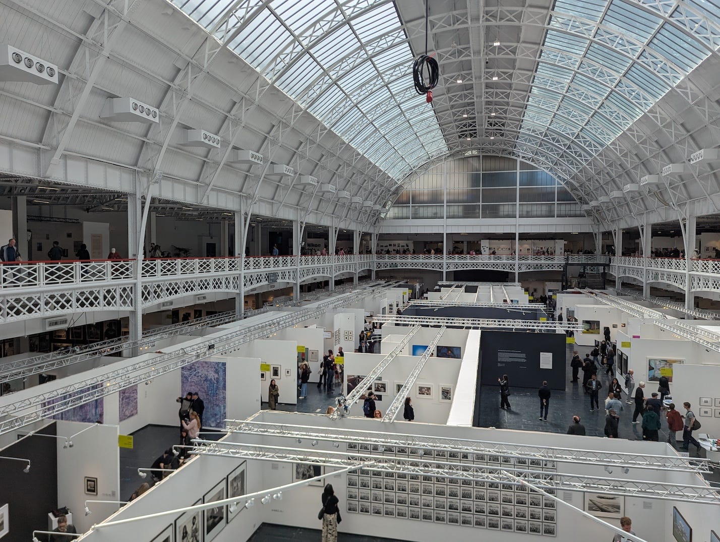 Wide interior shot of Olympia's grand hall with its sweeping glass and iron arched roof, filled with gallery stands and visitors browsing photography at Photo London.