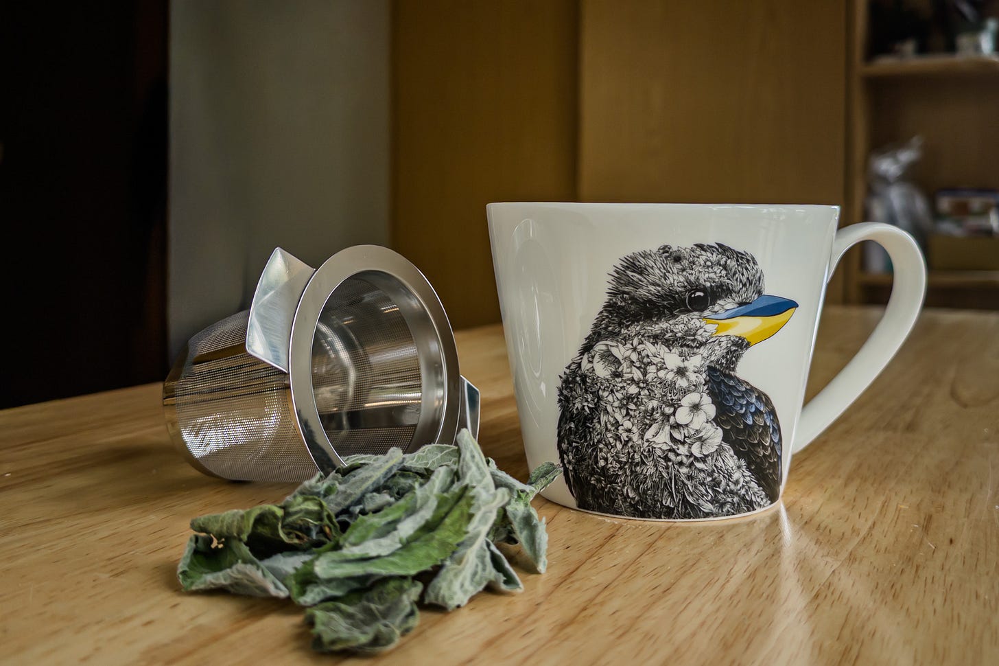 Auf einem Holztisch stehen ein metallenes Teesieb, eine weisse Tasse mit Eisvogel-Motiv und ein kleiner Haufen getrockneter Minzblätter. Die Szene zeigt die Vorbereitung für eine Tasse Minztee.