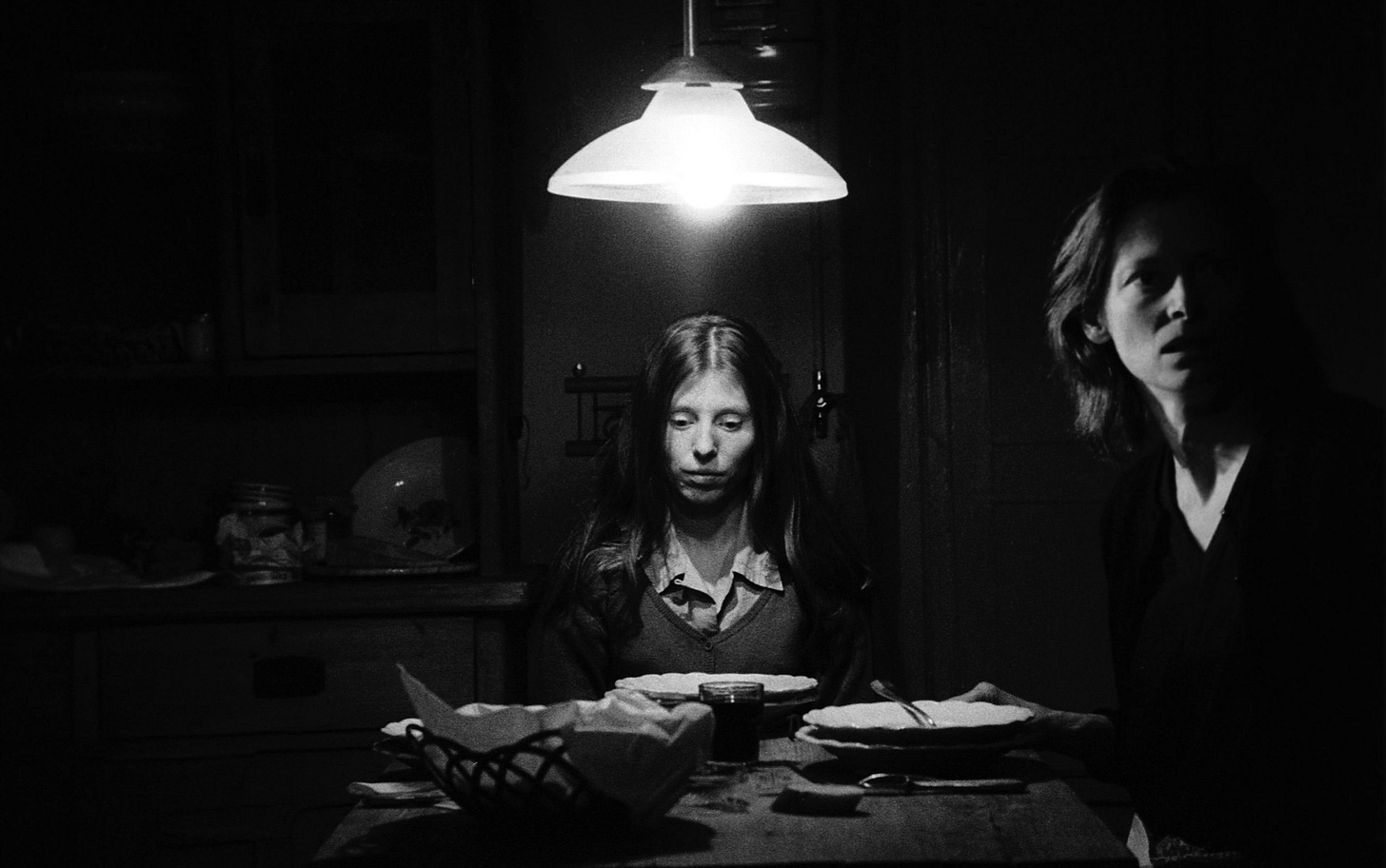 Henriette (Erika Bók) is sitting at the dinner table with her gaze lowered while her mother Camélia (Tilda Swinton) looks startled by something just off camera.