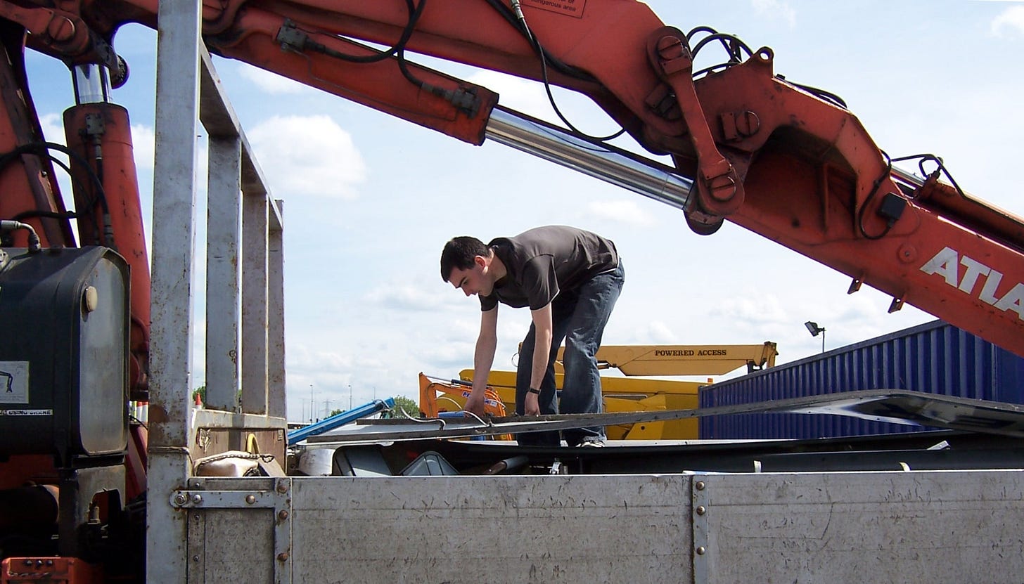 Photograph of a youthful version of this article's author standing on the back of a flat-bed truck, with a hydraulic crane arm above him, sorting through a pile of old road signs that are heaped up around him Photograph of a youthful version of this article's author standing on the back of a flat-bed truck, with a hydraulic crane arm above him, sorting through a pile of old road signs that are heaped up around him