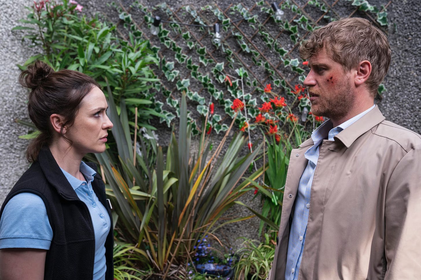 Janet, played by Róisín Gallagher and Seamus, played by Johnny Flynn look at each other, standing in front of a wall of plants.