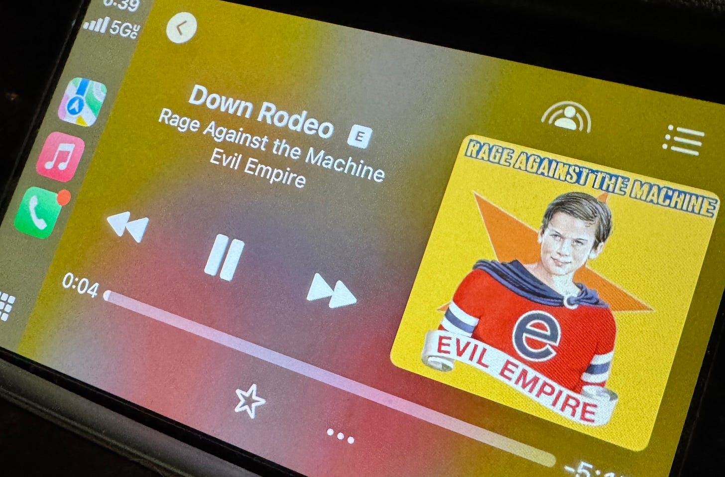 An Apple CarPlay screen displays the music player interface showing the song “Down Rodeo” by Rage Against the Machine from their album Evil Empire. The album artwork features a young boy in a red superhero-style outfit with a lowercase “e” on his chest and a banner reading “Evil Empire.” The background of the artwork is bright yellow with an orange star. The song is 4 seconds into playback, and the interface shows standard controls like pause, skip, and back. Icons for maps, music, and phone apps appear on the left. The device is connected to 5G.