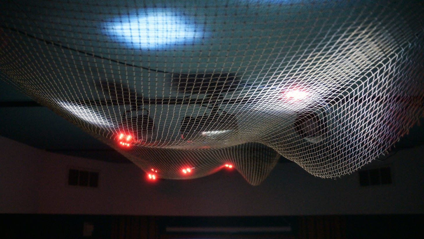 A suspended white net seen from below, red LED clusters and cool flashlight pools glowing across the mesh in a dark room A suspended white net seen from below, red LED clusters and cool flashlight pools glowing across the mesh in a dark room