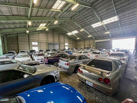 A collection of photos showing dozens of white, blue and green Nissan R34 Skyline GT-R sports cars crammed into a dimly-lit warehouse with gray walls and green support beams with a gravel floor.