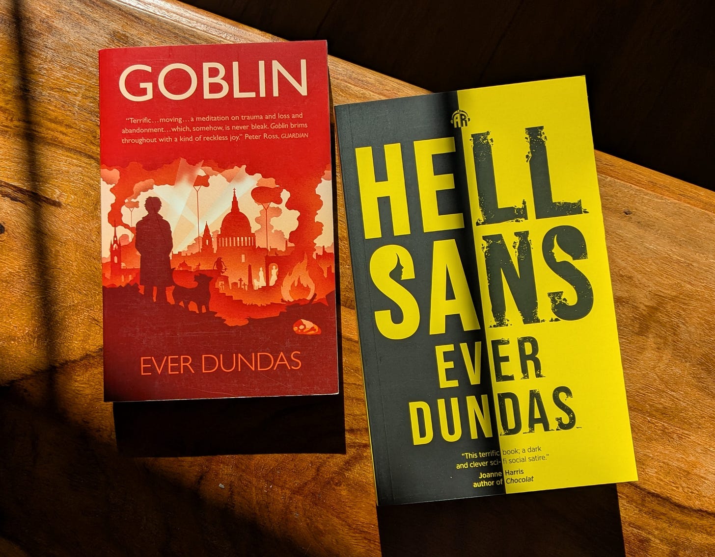  Two books by Ever Dundas on a table in bright sunlight, framed by heavy shadow. The first, Goblin, is a red cover featuring an illustration of bombed World War Two London, with a person and dog in silhouette watching from a distance.  The second is HellSans, which has a punk black and yellow cover. The title is in huge partially crumbling letters, with profiles of two women in the top curves of the ‘S’s in ‘Sans’.