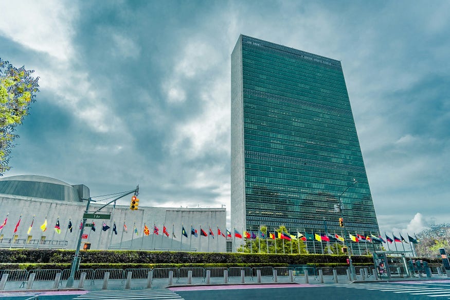 The United Nations building in New York City. Grey skies behind the skyscraper. An array of flags at ground level.