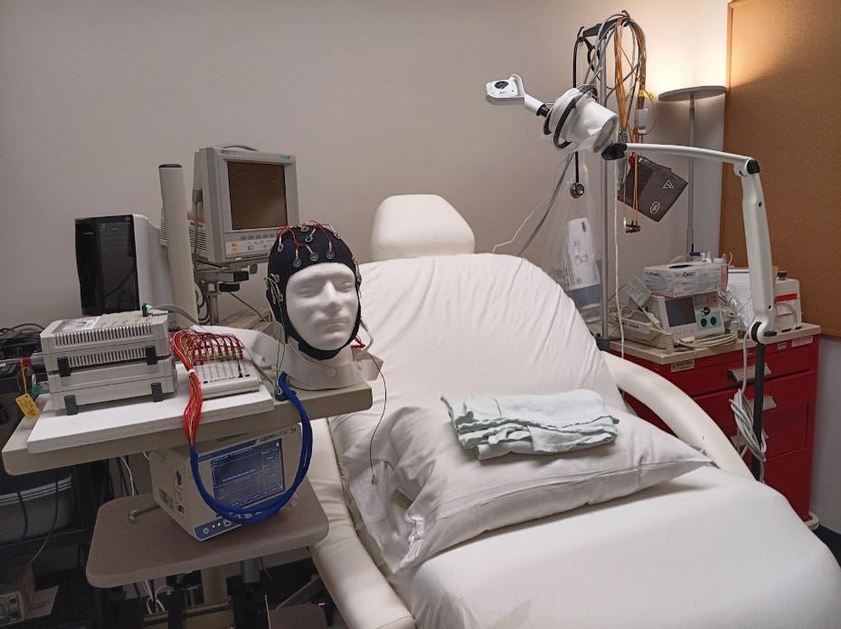 Medical room with an examination chair, EEG equipment, mannequin head with electrodes, and various medical devices.