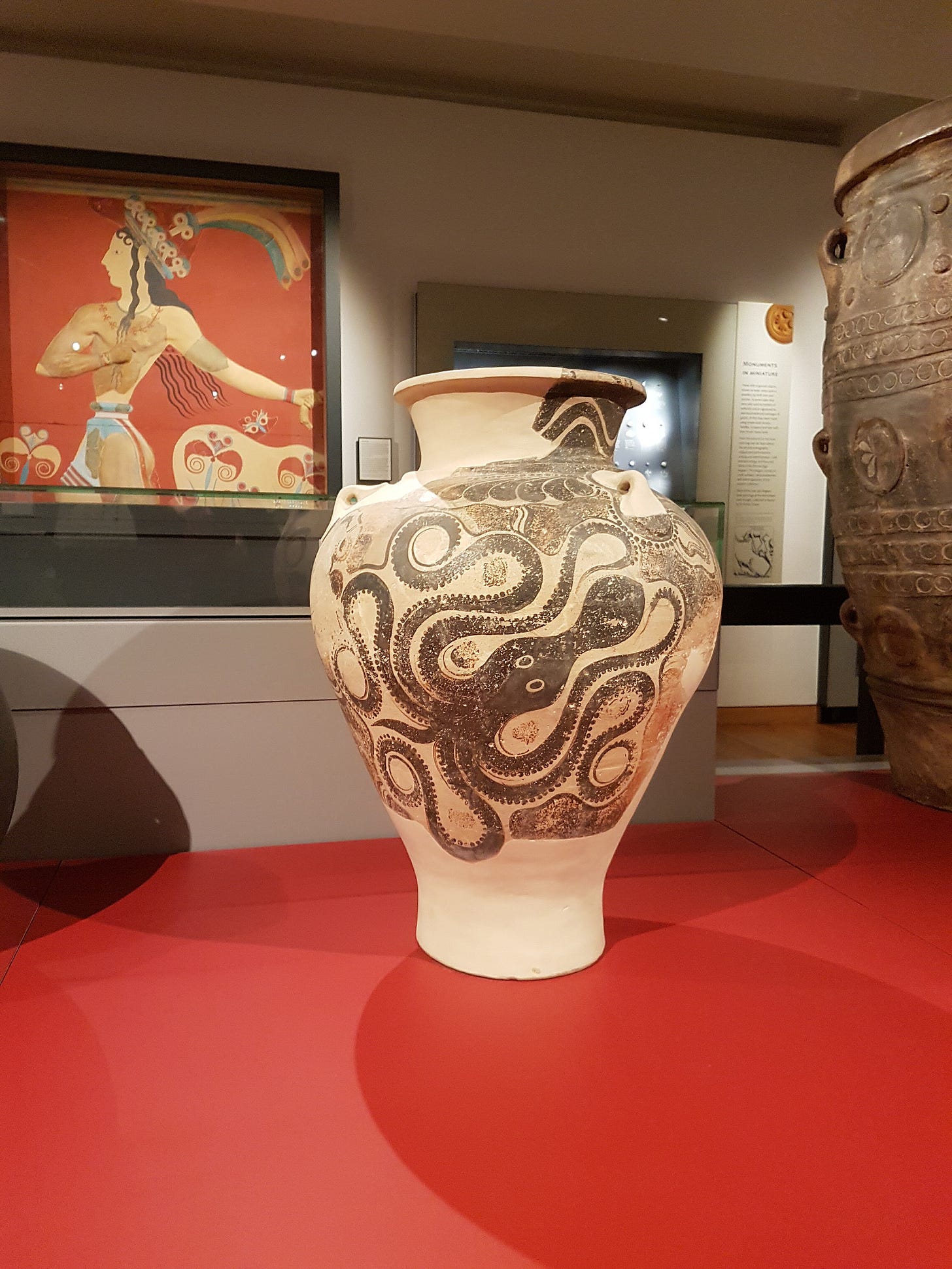 A tall Minoan pithos decorated with a swirling tentacled octopus sits on the floor in the middle of a museum, with the Minoan Prince of the Lilies fresco on the wall in the background.