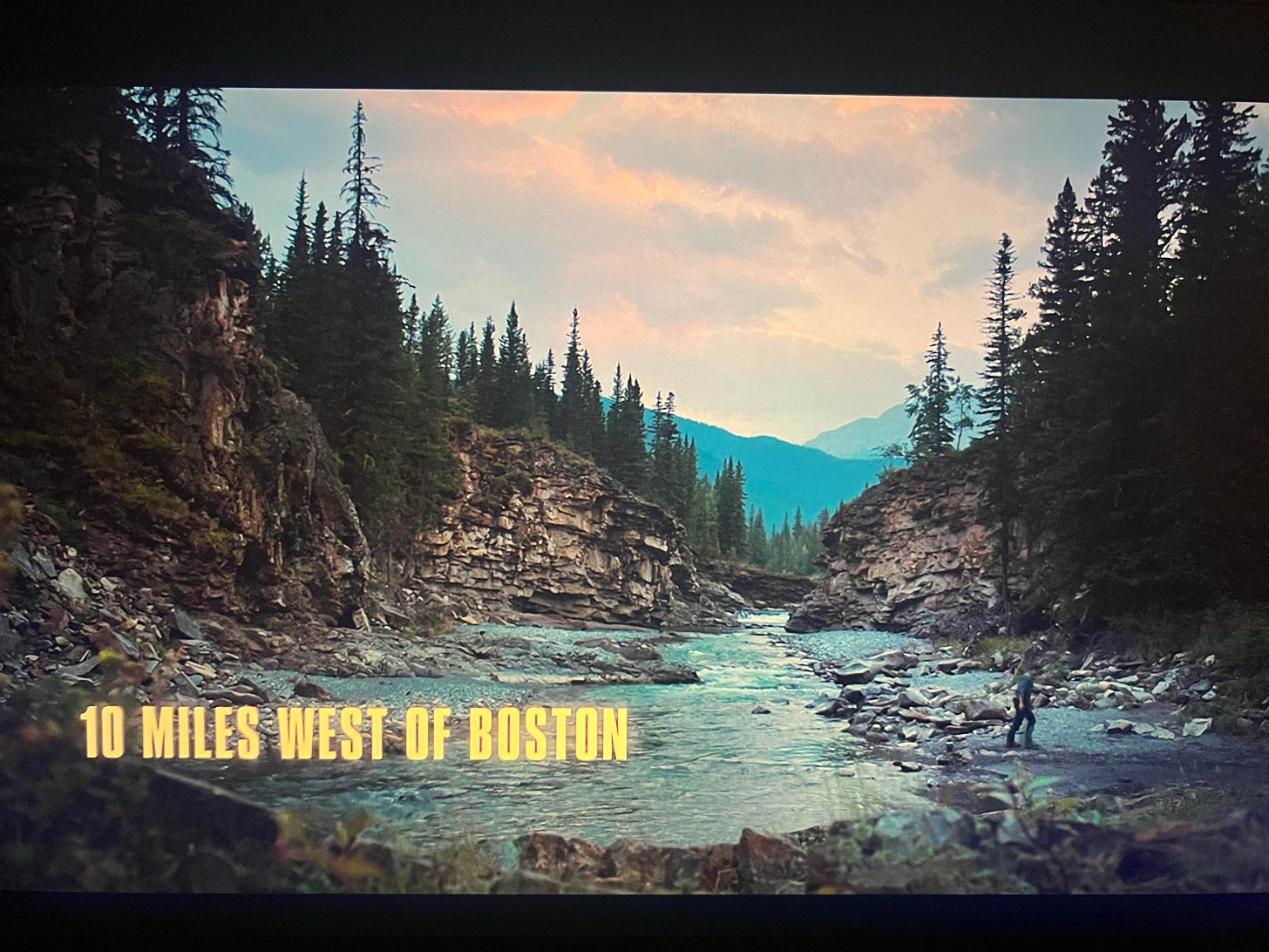 A screengrab from season 1, episode 2 of The Last of Us, featuring a craggy canyon, wide river, and stands of pine trees accompanied by bold yellow text that reads, "10 Miles West of Boston"