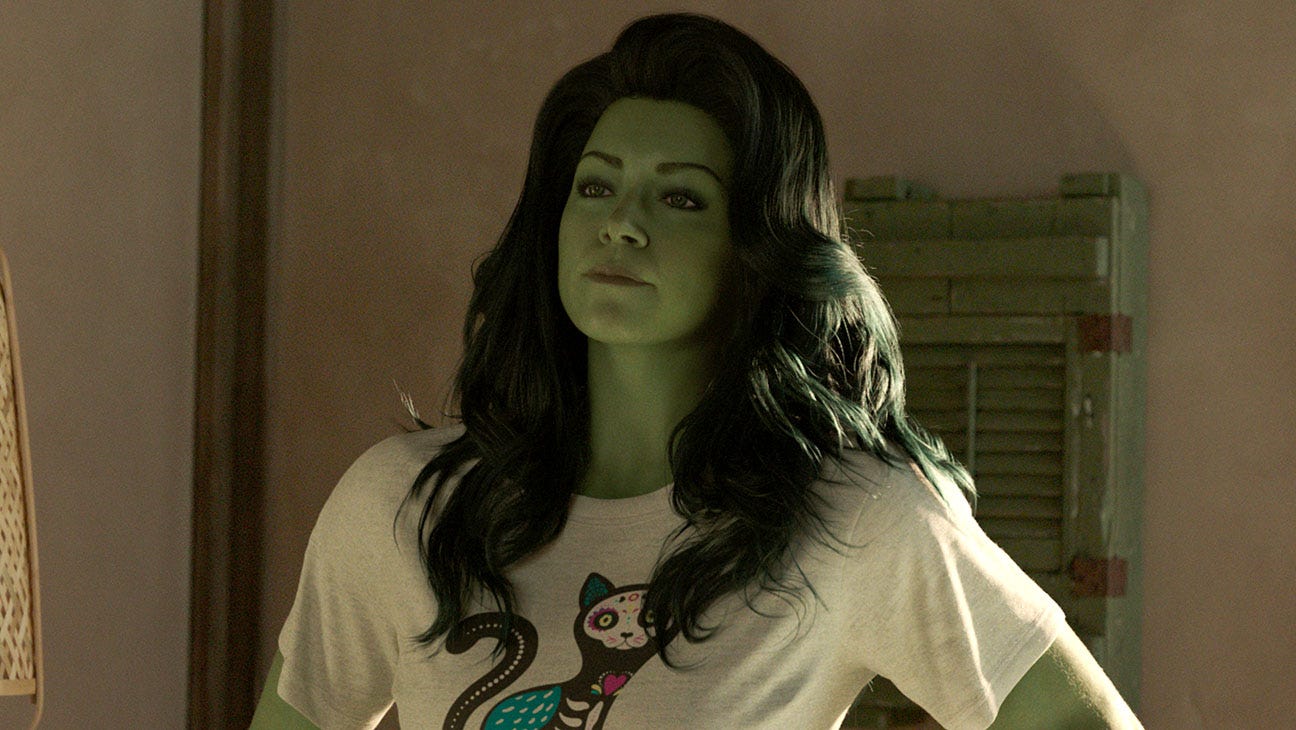 A imagem retrata a personagem Mulher-Hulk em um plano médio, do peito para cima, dentro de um ambiente com iluminação quente e suave. Ela apresenta sua característica pele verde, cabelos pretos longos e ondulados que caem sobre os ombros e uma expressão serena enquanto desvia o olhar ligeiramente para cima. A heroína veste uma camiseta branca de mangas curtas que traz no centro a estampa de um gato preto estilizado como uma caveira mexicana, adornado com detalhes coloridos em azul e rosa. Ao fundo, a composição mostra uma parede de tom claro e uma janela ou porta com venezianas de madeira pintadas de verde, completando o cenário residencial.