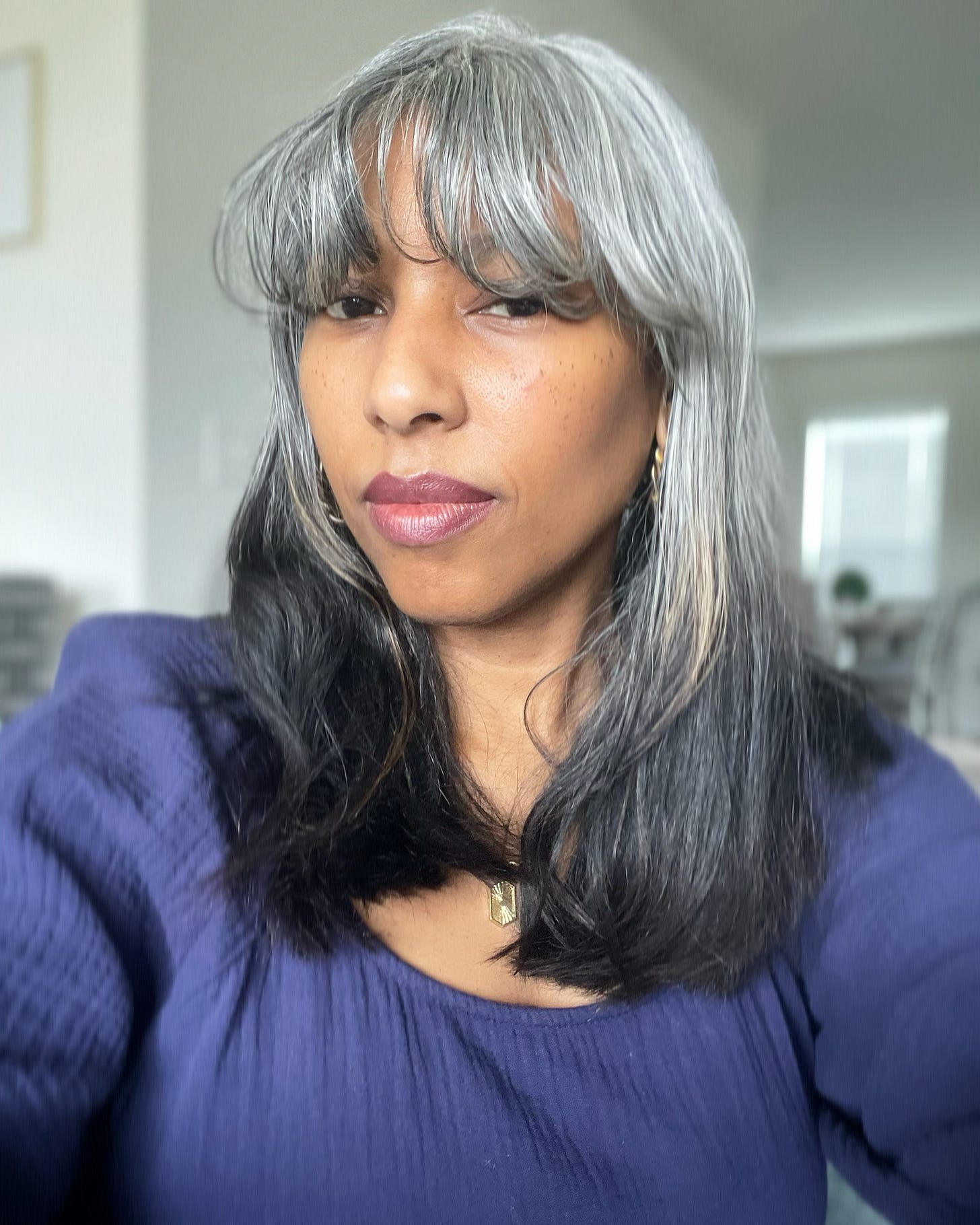 Khadijah wearing a dark blue cotton gauze top, not smiling. Her hair is past shoulder length and straight, with long bangs. The color is mostly white and gray, with the bottom 1/3 still black. A living room is blurred in the background. Her brown skin is somewhat pale.