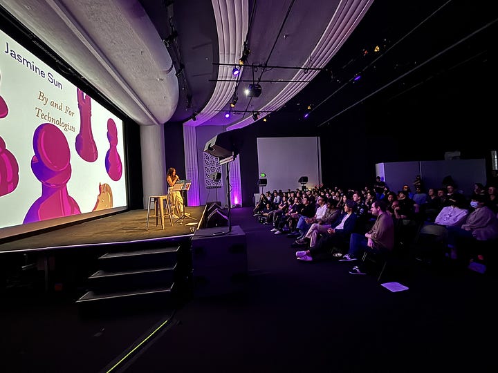 Kernel launch party in Gray Area event venue, including contributors in front of a screen, a seated audience at Jasmine's talk, magazines on a table, and various people mingling under purple lighting
