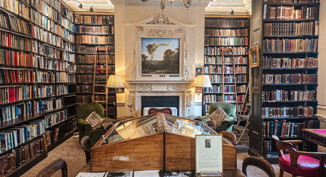 A room inside Bromley House Library with bookshelves, tables, chairs, and a white wall with artwork and a fireplace next to it