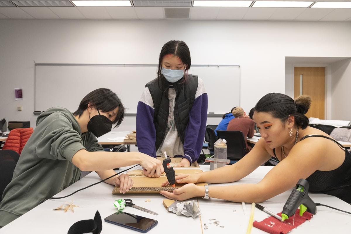 three students work together on a cardboard prototype in a classroom, with hot glue and screws and scissors and other supplies strewn around the table three students work together on a cardboard prototype in a classroom, with hot glue and screws and scissors and other supplies strewn around the table