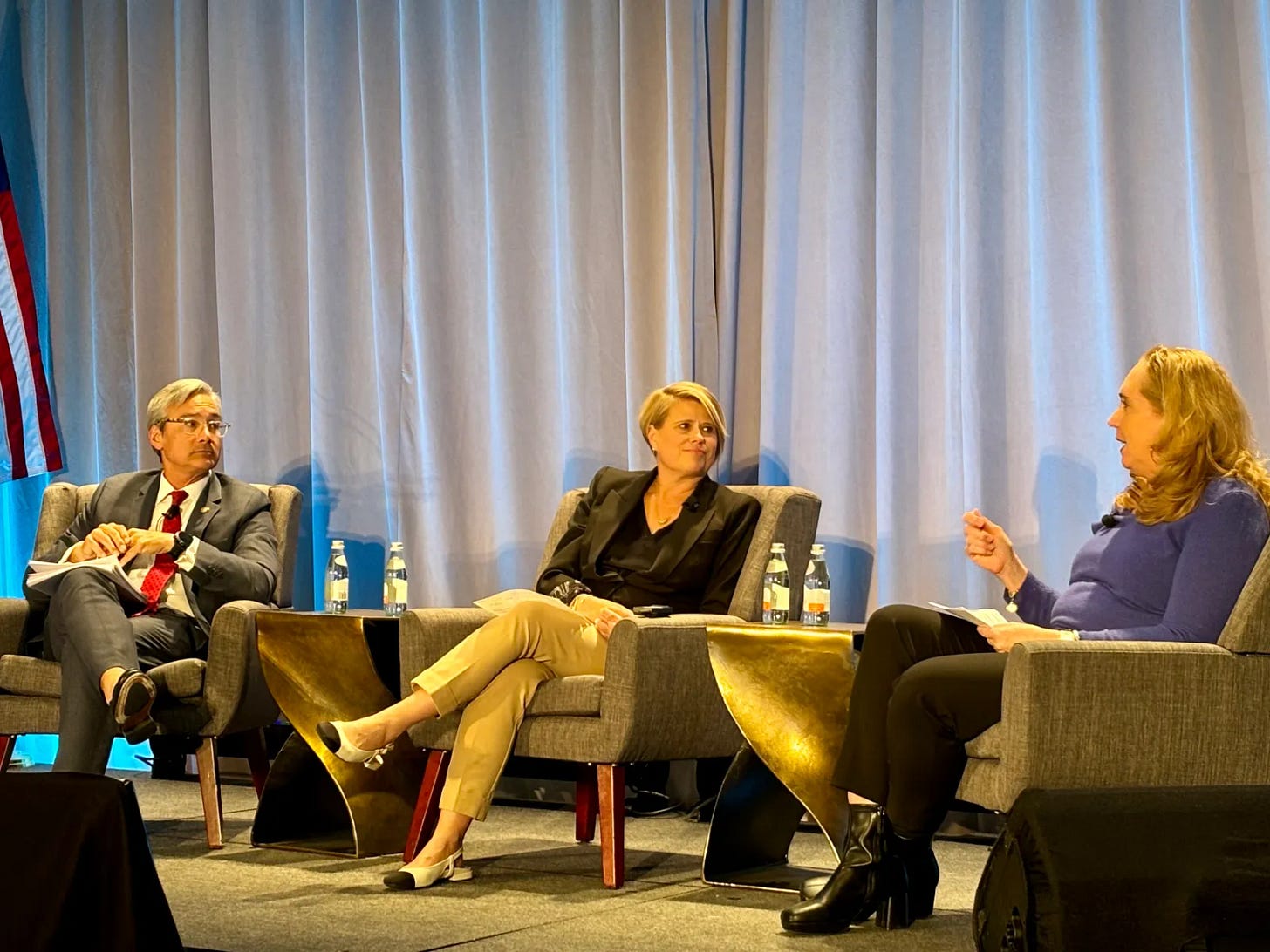 Three people sit in chairs on a stage. A curtain and US flag are behind them Three people sit in chairs on a stage. A curtain and US flag are behind them