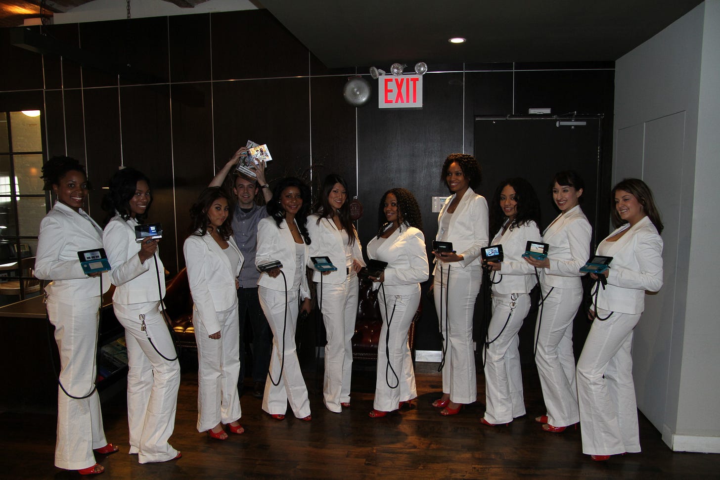 Photo of 10 women in white pantsuits holding Nintendo handheld systems that are tethered to their outfits. Photo of 10 women in white pantsuits holding Nintendo handheld systems that are tethered to their outfits.