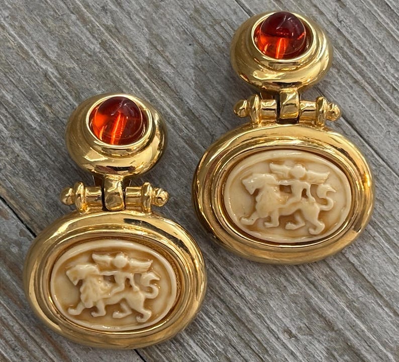 May include: A pair of gold-toned clip-on earrings. Each earring features a round, orange-red cabochon at the top and an oval pendant with a cream-colored relief of two mythical creatures. The earrings have a hinged design.
