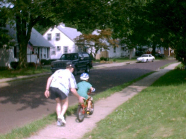 This may contain: a man pushing a boy on a bike down a sidewalk next to a street with houses