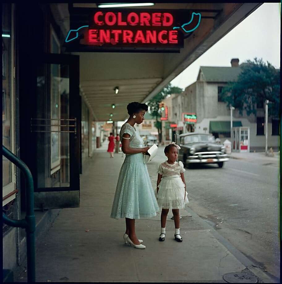 Photo by Gordon Parks, taken in his photographic journey published as part of "Segregation Story" as he travelled in Alabama during the late 1950s. A Black woman and Black girl are both elegantly dressed outside a store. Above them, a neon sign points towards the entrance saying "COLORED ENTRANCE".