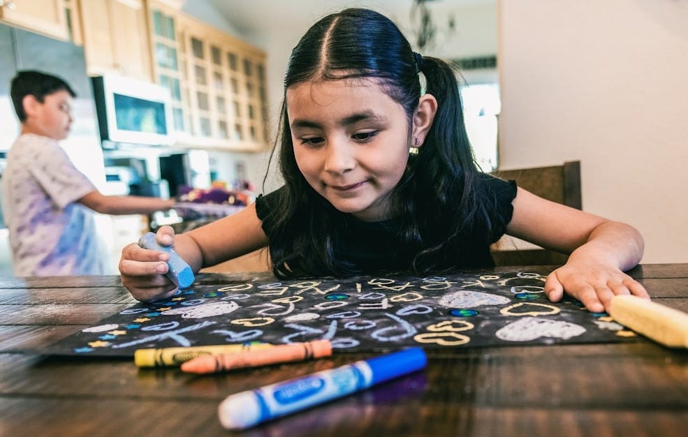 a little girl sitting at a table with crayons and markers