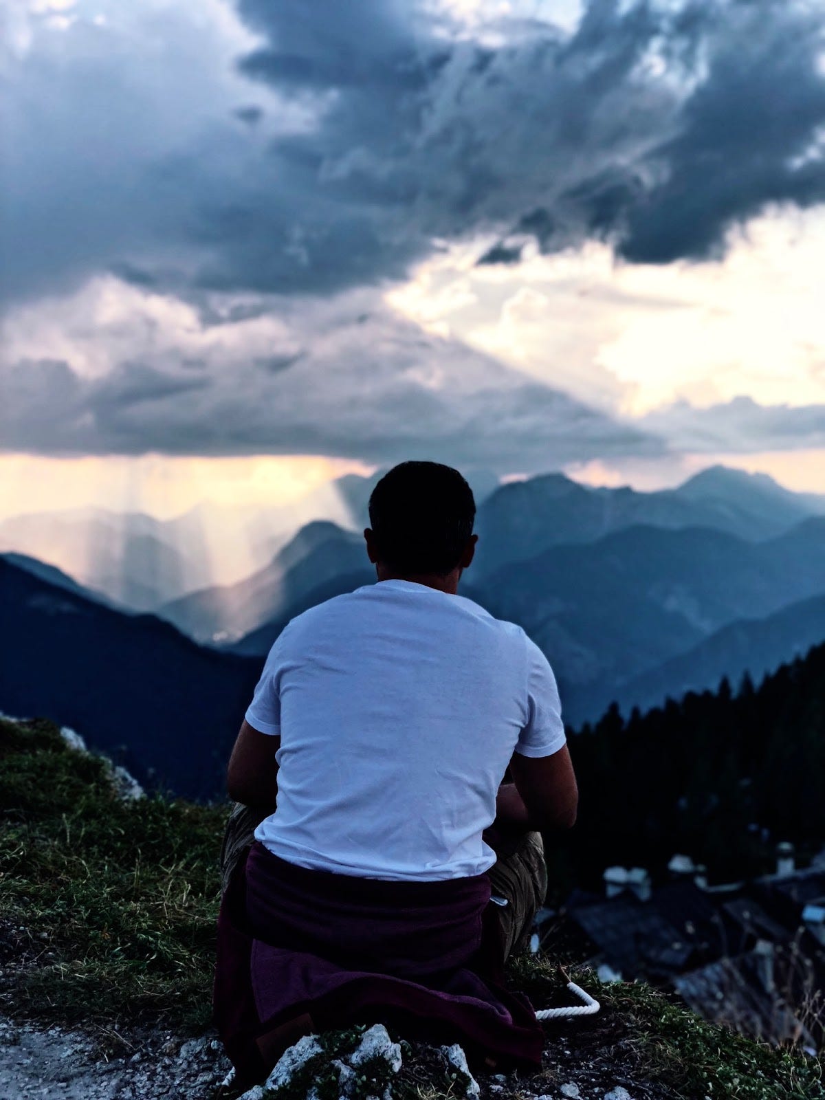 Un uomo di spalle, seduto su un prato, osserva le montagne in lontananza al tramonto