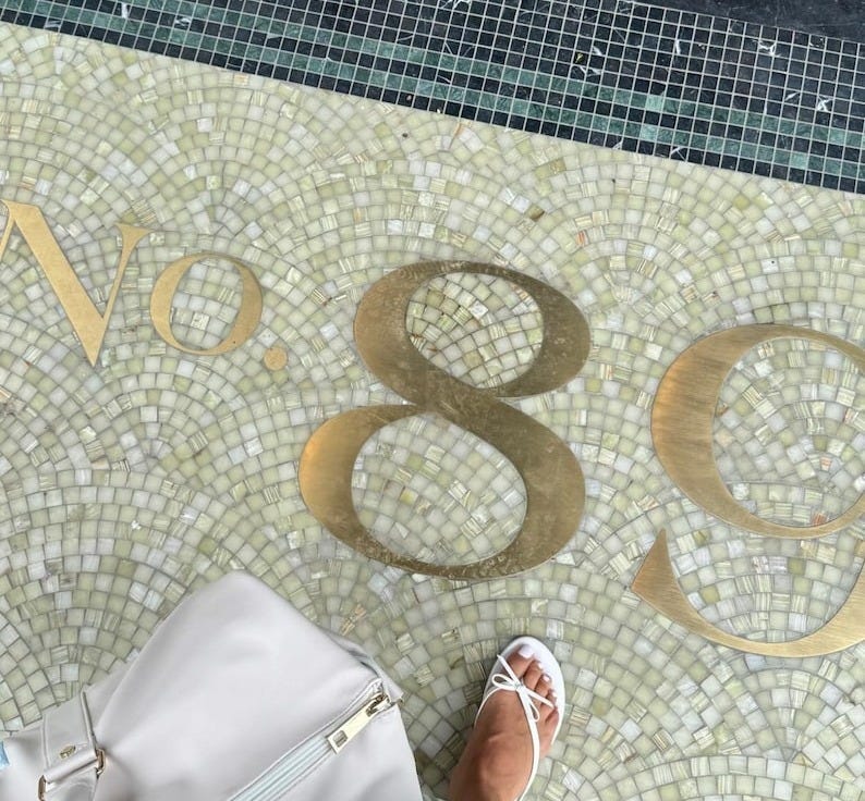 May include: A mosaic tile floor with the number "89" in gold lettering. A white bag and a person's foot in white sandals are visible. The door frame is dark wood. May include: A mosaic tile floor with the number "89" in gold lettering. A white bag and a person's foot in white sandals are visible. The door frame is dark wood.