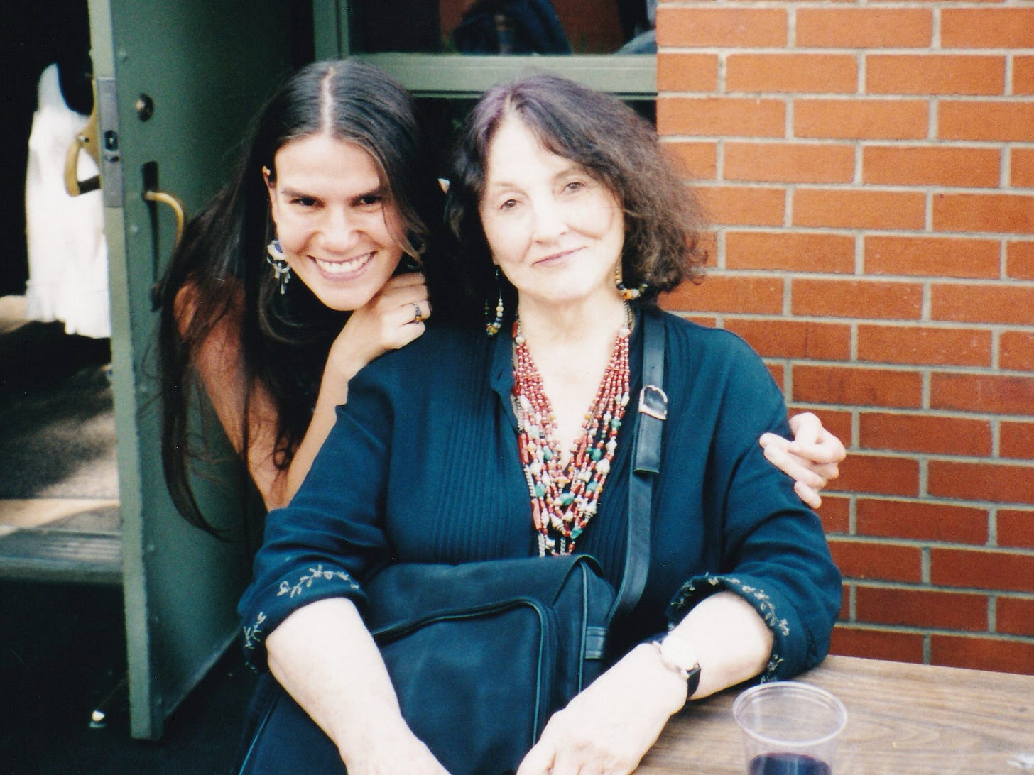 A woman (Alexandra Hidalgo) is standing and she leans forward to put her arms around a woman who (Bobbie Lousie Hawkins) is sitting at a table. They smile at the camera. They are outside with brick walls behind them. It's afternoon. A woman (Alexandra Hidalgo) is standing and she leans forward to put her arms around a woman who (Bobbie Lousie Hawkins) is sitting at a table. They smile at the camera. They are outside with brick walls behind them. It's afternoon.