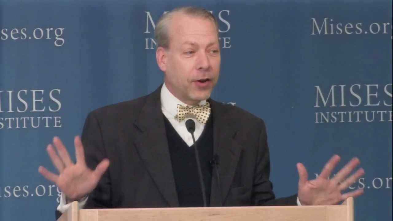 Jeffrey Tucker in a polka dot bow tie gestures with both hands at a Mises Institute podium. Jeffrey Tucker in a polka dot bow tie gestures with both hands at a Mises Institute podium.