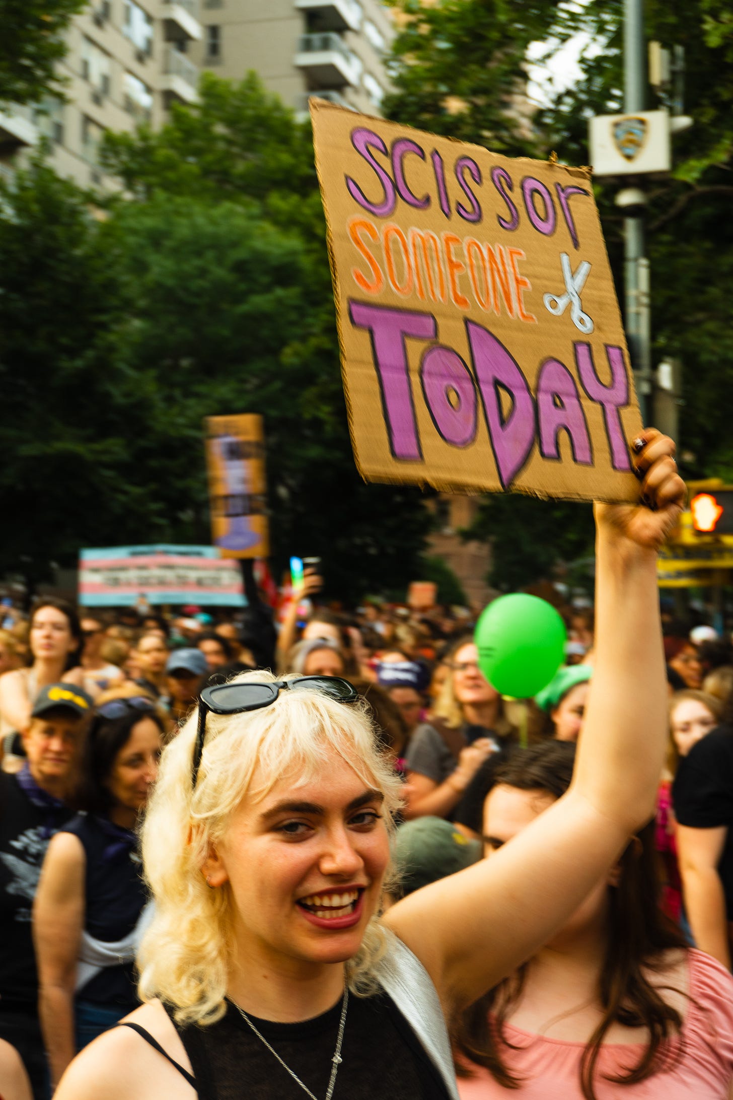 A queer person smiles at the camera and holds up a sign that reads: "scissor someone today"